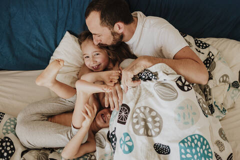 Vater küsst Tochter auf die Stirn, während Sohn lachend auf dem Bett zu Hause, lizenzfreies Stockfoto