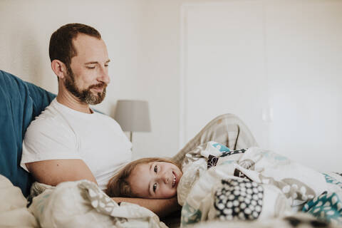 Lächelnde Tochter auf dem Schoß des Vaters auf dem Bett zu Hause liegend, lizenzfreies Stockfoto