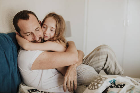 Daughter leaning on father while hugging on bed at home - GMLF01007