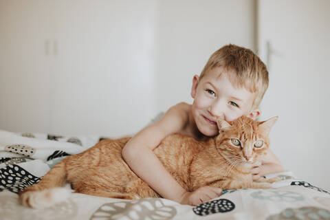 Lächelnder Junge, der sich im Schlafzimmer zu Hause an eine rothaarige Katze lehnt, lizenzfreies Stockfoto