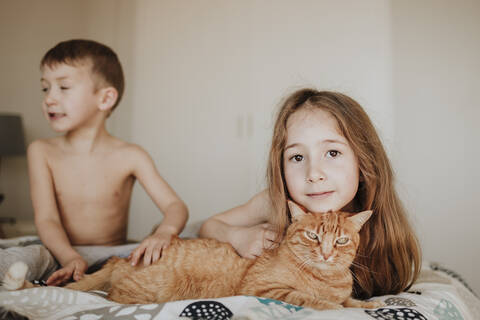 Girl leaning on cat while brother looking away in bedroom at home stock photo