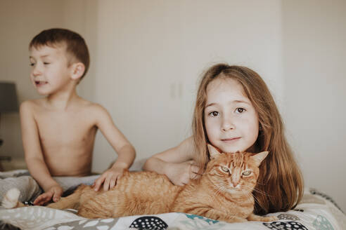 Girl leaning on cat while brother looking away in bedroom at home - GMLF01012