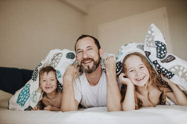 Smiling father with daughter and son lying on bed under blanket in bedroom at home - GMLF01014