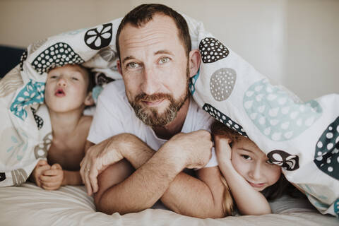 Verspielter Junge mit Vater und Schwester unter einer Decke auf dem Bett zu Hause, lizenzfreies Stockfoto