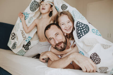 Cheerful daughter with father while brother looking away under blanket on bed at home stock photo