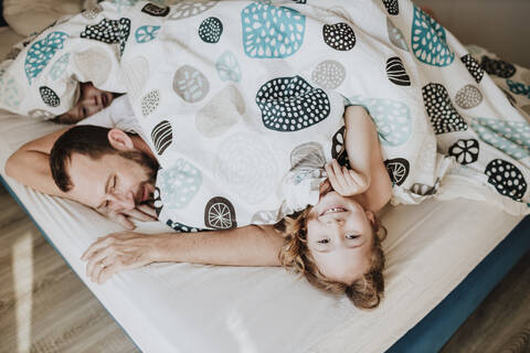Lächelndes Mädchen mit Vater und Bruder auf dem Bett liegend unter einer Decke zu Hause, lizenzfreies Stockfoto