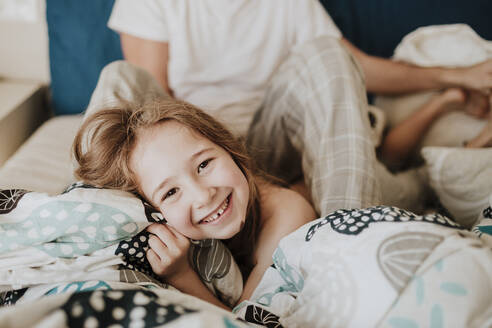Cheerful girl lying on bed with father sitting in background at home - GMLF01019