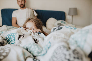 Mädchen versteckt sich unter einer Decke, während der Vater zu Hause auf dem Bett zusieht - GMLF01020