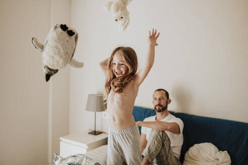 Fröhliches Mädchen wirft Plüschtiere, während der Vater zu Hause auf dem Bett zusieht - GMLF01021