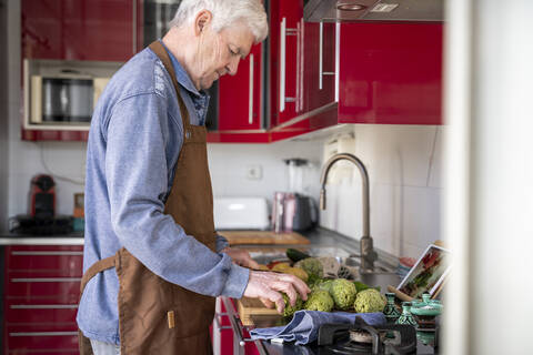 Senior man in apron cutting artichokes while doing video call with daughter on digital tablet at home stock photo