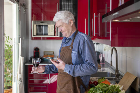 Senior man holding glass of wine while using digital tablet at kitchen stock photo