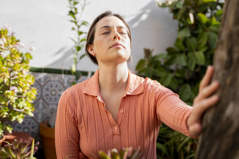 Young woman with eyes closed touching tree trunk in garden - AFVF08262