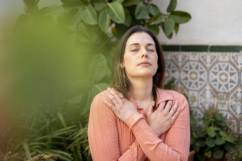 Beautiful young woman with eyes closed hugging self in garden - AFVF08275