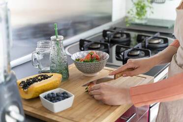 Young woman slicing strawberry with knife on cutting board in kitchen - AFVF08281