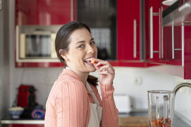 Cheerful woman eating slice of strawberry in kitchen - AFVF08284