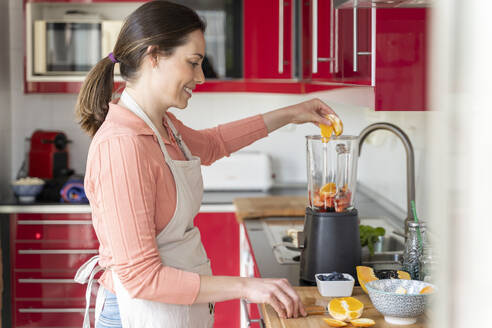 Smiling young woman preparing healthy fruit smoothie in kitchen at home - AFVF08287