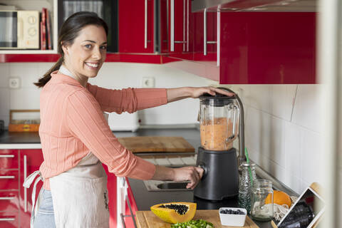 Lächelnde junge Frau bei der Zubereitung eines gesunden Fruchtsmoothies in der Küche zu Hause, lizenzfreies Stockfoto