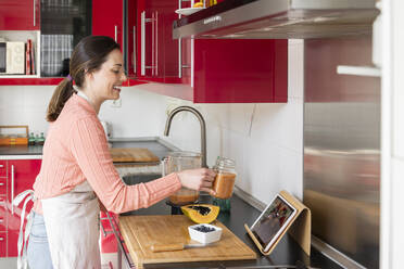 Smiling woman showing smoothie jar to friends on video call while standing in kitchen at home - AFVF08300