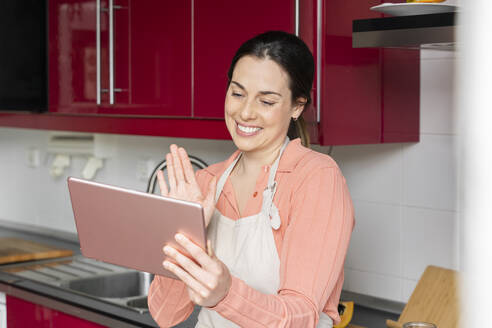 Beautiful woman waving hand during video call on digital tablet in kitchen at home - AFVF08304