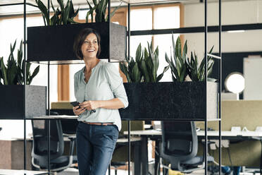 Lächelnde Geschäftsfrau mit Smartphone, die wegschaut, während sie im Büro steht - JOSEF03620