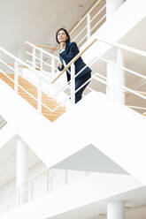 Female entrepreneur with hand on chin contemplating while standing over staircase - JOSEF03630