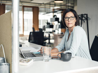 Selbstbewusste Unternehmerin mit Laptop und Blick auf den Schreibtisch im Büro - JOSEF03633