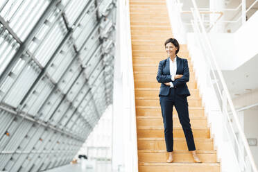 Smiling businesswoman with arms crossed standing on steps in corridor - JOSEF03634