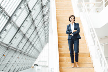 Confident businesswoman with arms crossed standing on staircase in corridor - JOSEF03635