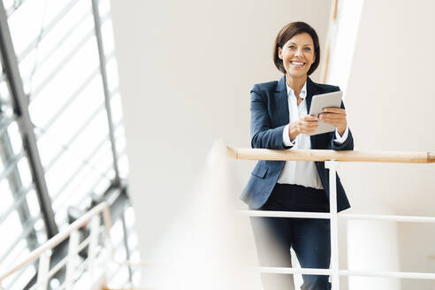 Happy businesswoman with digital tablet leaning on railing at corridor - JOSEF03641