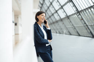 Businesswoman talking on smart phone while leaning on wall in corridor - JOSEF03643
