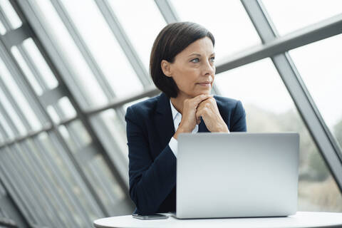 Nachdenkliche Geschäftsfrau mit Hand am Kinn neben dem Laptop am Schreibtisch im Büro, lizenzfreies Stockfoto