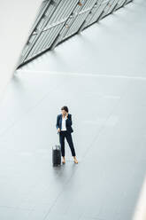 Businesswoman with suitcase talking on smart phone while standing at corridor - JOSEF03646