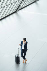 Female entrepreneur with suitcase talking on mobile phone while standing at corridor - JOSEF03647