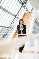 Smiling female entrepreneur with hand on chin leaning over railing in corridor - JOSEF03653