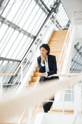 Businesswoman with hand on chin leaning over railing in corridor - JOSEF03654