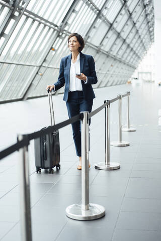 Female professional with smart phone and suitcase walking in corridor stock photo