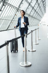 Female professional with smart phone and suitcase walking in corridor - JOSEF03663