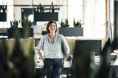Smiling businesswoman with hands in pockets leaning on desk at office - JOSEF03664