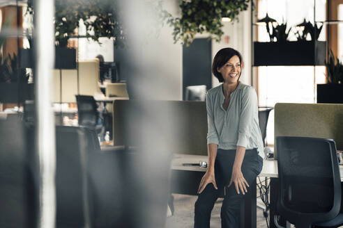 Happy businesswoman looking away while leaning on desk in office - JOSEF03665