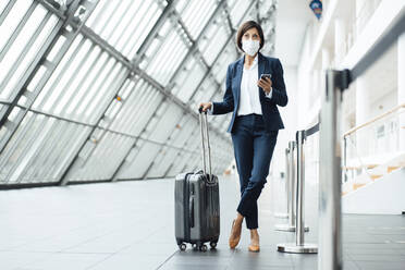 Businesswoman with smart phone and suitcase standing in corridor during COVID-19 - JOSEF03675