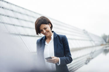 Lächelnde Geschäftsfrau mit Textnachrichten auf einem Smartphone vor einem Bürogebäude - JOSEF03685