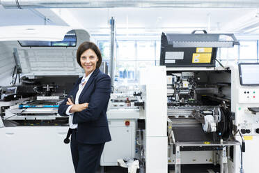 Smiling female entrepreneur with arms crossed against machinery in factory - JOSEF03692