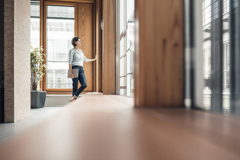 Businesswoman with laptop looking through window at office - JOSEF03712