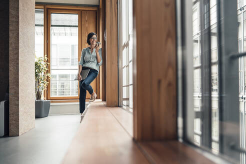 Mature businesswoman with smart phone standing by window at office - JOSEF03714