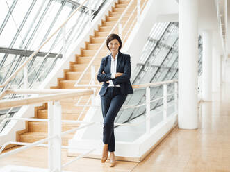 Smiling confident businesswoman with arms crossed by railing in corridor - JOSEF03739