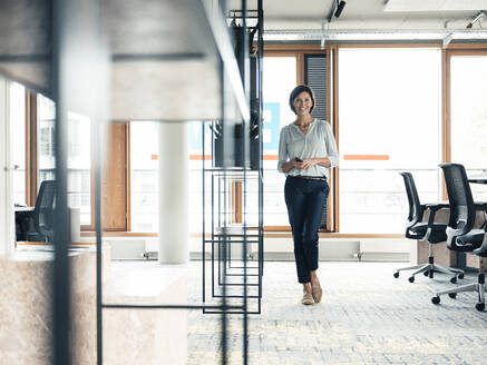 Happy female entrepreneur with smart phone standing at office - JOSEF03753