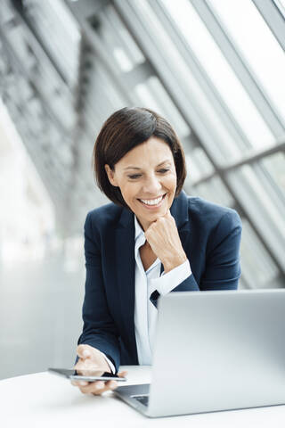 Smiling businesswoman with hand on chin working on laptop in corridor stock photo