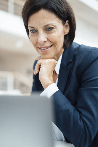 Smiling businessman with hand on chin using laptop at office stock photo