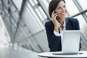 Businesswoman smiling while talking on smart phone at desk in corridor - JOSEF03777