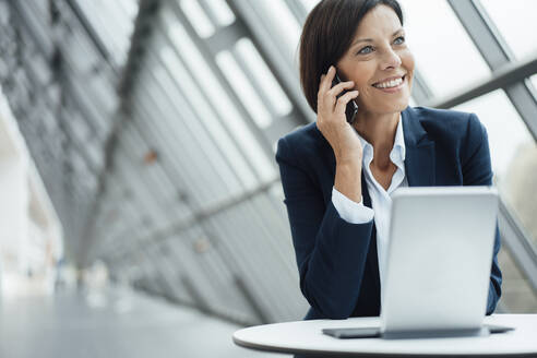 Geschäftsfrau lächelt, während sie am Schreibtisch im Korridor mit einem Smartphone spricht - JOSEF03777